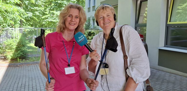 Joanna Rutkowska trzymająca w dłoniach kije do nordic walking oraz Anna Rębas. W tle fragment budynku i zielone drzewa