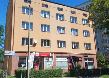 Gdynia. Czterokondygnacyjna kamienica o beżowo-pomarańczowej elewacji. Na parterze budynku widoczne są lokale usługowe (m.in. ubezpieczenia i fryzjer), a przed nim zadbany żywopłot i ulica w słoneczny dzień
