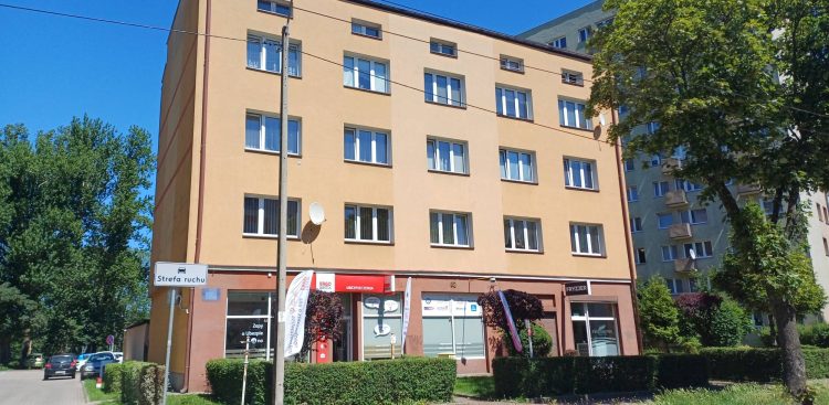 Gdynia. Czterokondygnacyjna kamienica o beżowo-pomarańczowej elewacji. Na parterze budynku widoczne są lokale usługowe (m.in. ubezpieczenia i fryzjer), a przed nim zadbany żywopłot i ulica w słoneczny dzień