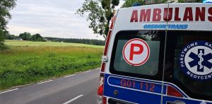 Zbliżenie na tylną część białej karetki pogotowia marki Gregor Trans zaparkowanej na poboczu drogi obok zielonego pola i drzew pod zachmurzonym niebem. Na drzwiach widoczny jest czerwony znak „P” (zespół podstawowy), napis „GREGOR TRANS”, numer boczny „G01 112” oraz logo Państwowego Ratownictwa Medycznego z Gwiazdą Życia. Wóz jest na miejscu wypadku, w tle widać odległe zabudowania. Zdjęcie jest ilustracją do wypadku na budowie obwodnicy Kartuz, gdzie mężczyzna pracujący w wykopie został przysypany ziemią