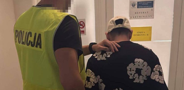 Policjant w odblaskowej kamizelce z napisem "POLICJA" prowadzi zatrzymanego mężczyznę w czarnej wzorzystej koszulce i białej czapce z daszkiem do gabinetu z tabliczką "REFERAT KRYMINALNY"