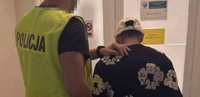 Policjant w odblaskowej kamizelce z napisem "POLICJA" prowadzi zatrzymanego mężczyznę w czarnej wzorzystej koszulce i białej czapce z daszkiem do gabinetu z tabliczką "REFERAT KRYMINALNY"