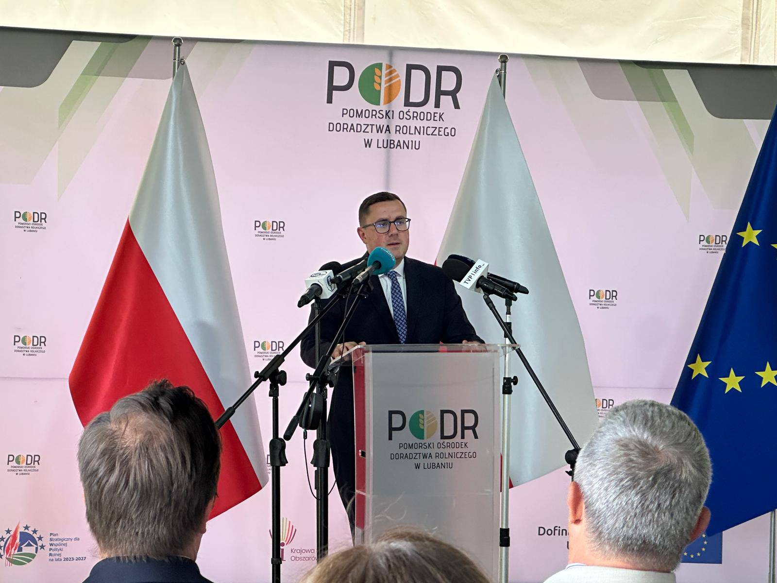 Mężczyzna w okularach i garniturze przemawia za mównicą z logo PODR w Lubaniu, otoczony flagami Polski i Unii Europejskiej oraz mikrofonami dziennikarzy. Konferencja prasowa w Pomorskim Ośrodku Doradztwa Rolniczego w Lubaniu poświęcona budowie "Wyspy Energetycznej Lubań"