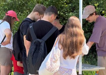 Grupa ludzi stojących pod białym namiotem w plenerze, pochylających się nad stolikiem z mapami i materiałami konsultacyjnymi w otoczeniu zieleni. Konsultacje społeczne masterplanu Gdańsk Nowe Południe
