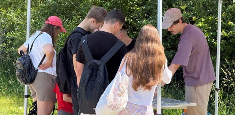 Grupa ludzi stojących pod białym namiotem w plenerze, pochylających się nad stolikiem z mapami i materiałami konsultacyjnymi w otoczeniu zieleni. Konsultacje społeczne masterplanu Gdańsk Nowe Południe