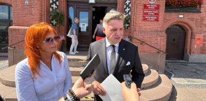 Poseł Michał Kowalski w czarnym garniturze i radna Marzena Gmińska stoją na schodach przed Urzędem Miejskim w Słupsku, udzielając wypowiedzi dziennikarzom trzymającym dyktafony i telefony. Chodzi o incydenty z udziałem obcokrajowców