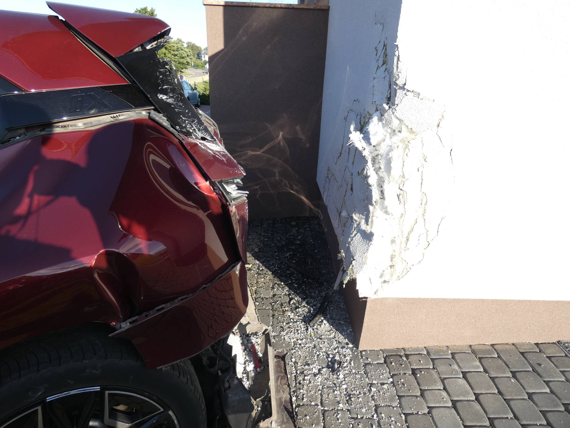 Wypadek w Niedamowie. Zbliżenie na miejsce uderzenia: mocno wgnieciony narożnik i rozbity tył bordowego samochodu oraz głęboko skruszona, pęknięta biała elewacja budynku z odsłoniętą warstwą ocieplenia. Na kostce brukowej leżą kawałki tynku i potłuczone szkło