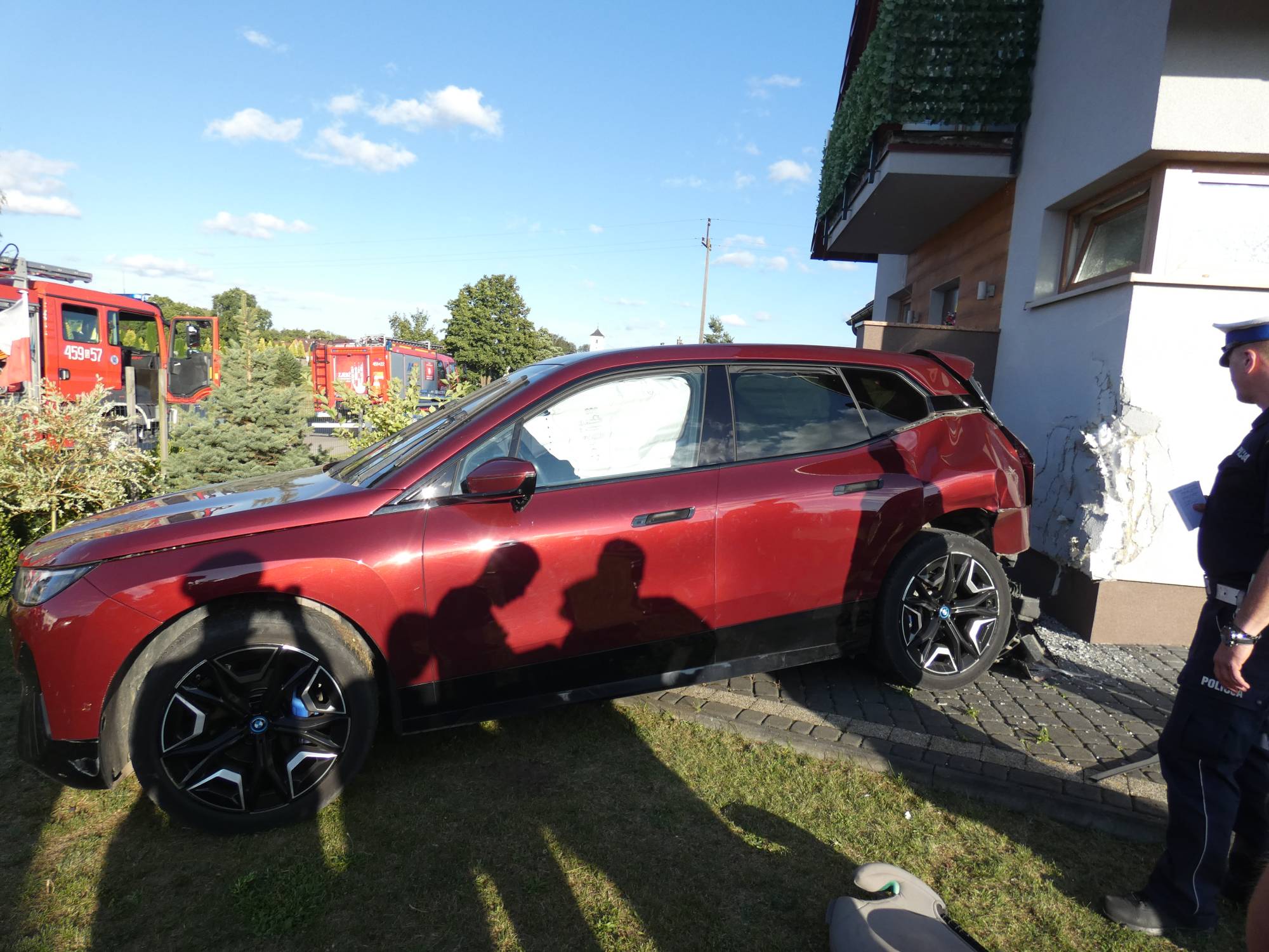 Bordowy samochód osobowy marki BMW z wystrzelonymi kurtynami powietrznymi, wbity tyłem w narożnik elewacji domu jednorodzinnego. Obok pojazdu stoi policjant sporządzający notatkę, na trawniku widać fotelik dziecięcy, a w tle stoją wozy strażackie