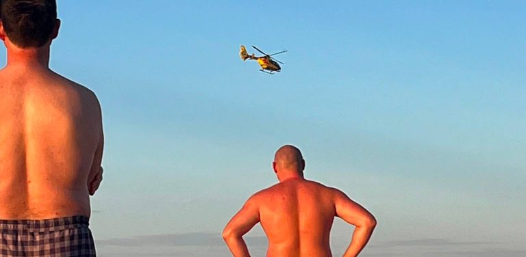 Akcja ratunkowa w Ustce. Widok od tyłu na dwóch mężczyzn stojących na plaży i patrzących w górę, gdzie nad nimi nisko nad ziemią leci żółto-czerwony śmigłowiec Lotniczego Pogotowia Ratunkowego na tle jasnoniebieskiego nieba