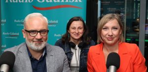 Na zdjęciu znajdują się trzy osoby patrzące wprost w kamerę - dwie kobiety po prawej i mężczyzna po lewej. Przed nimi znajdują się mikrofony.
