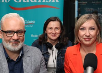 Na zdjęciu znajdują się trzy osoby patrzące wprost w kamerę - dwie kobiety po prawej i mężczyzna po lewej. Przed nimi znajdują się mikrofony.