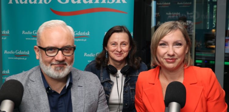 Na zdjęciu znajdują się trzy osoby patrzące wprost w kamerę - dwie kobiety po prawej i mężczyzna po lewej. Przed nimi znajdują się mikrofony.