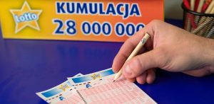 Dłoń skreślająca ołówkiem liczby na blankiecie gry Lotto. W tle widoczny jest żółty banner z logo Lotto i napisem „KUMULACJA 28 000 000”