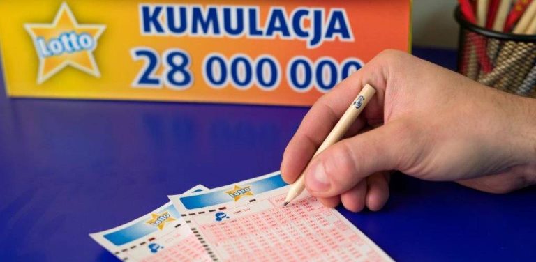 Dłoń skreślająca ołówkiem liczby na blankiecie gry Lotto. W tle widoczny jest żółty banner z logo Lotto i napisem „KUMULACJA 28 000 000”