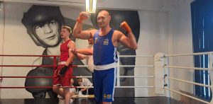 Marcin Piejek z KS Sako Gdańsk po zwycięskiej walce w czasie III Memoriału Jana Biangi w klubie bokserskim Brzostek Top Team w Gdańsku. Bokser w niebieskim stroju sportowym i z owiniętymi na pomarańczowo dłońmi stoi w ringu z uniesionymi rękami w geście zwycięstwa. W tle widoczny jest jego rywal w czerwonym stroju oraz malowidło ścienne przedstawiające postać boksera Jana Biangi