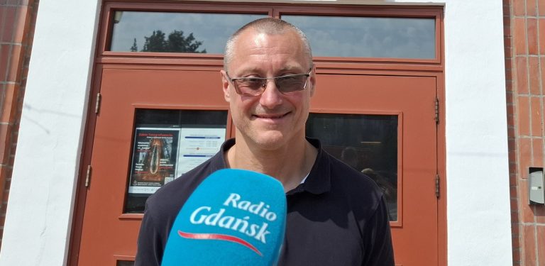 Uśmiechnięty, krótkowłosy mężczyzna w okularach i ciemnej koszulce polo, stojący przed brązowymi drzwiami budynku. Przed nim znajduje się niebieski mikrofon z logo Radia Gdańsk. To Wicedyrektor Narodowego Muzeum Morskiego w Gdańsku, Marcin Westphal, który zapowiadał nową wystawę poświęconą polskim transatlantykom m.s. Piłsudski oraz m.s. Batory