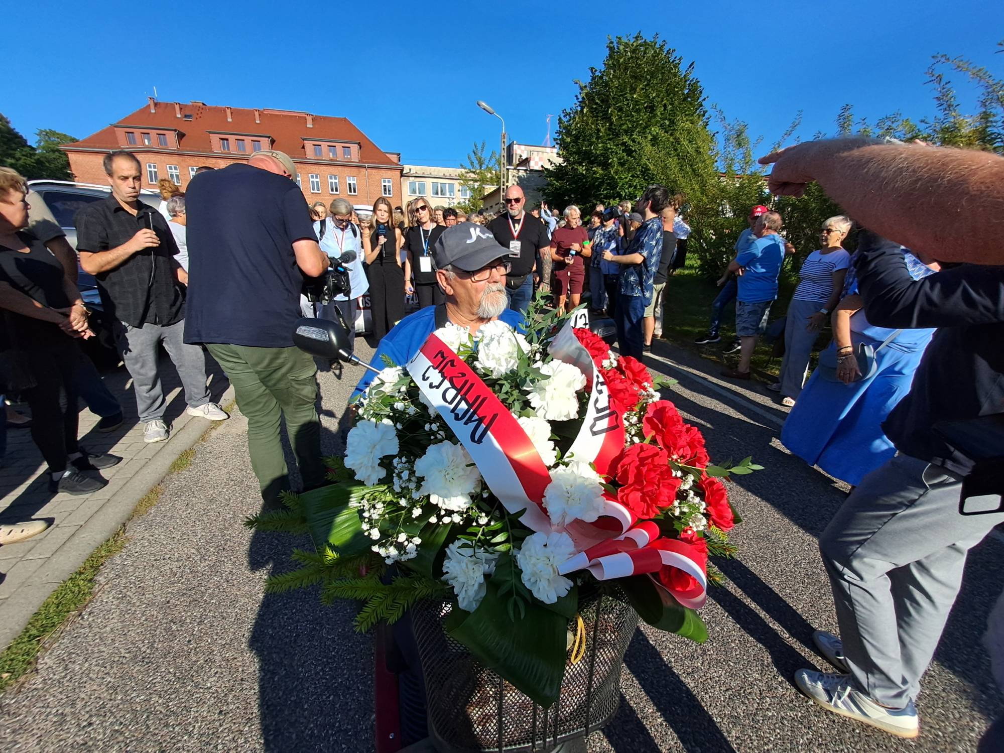 Marsz milczenia w obronie szpitala w Miastku. Mężczyzna w czapce z daszkiem i okularach przewozi w bagażniku inwalidzkiego wózka elektrycznego żałobną wiązankę kwiatów z biało-czerwoną szarfą z napisem „Marsz ciszy”. Wokół niego gromadzą się uczestnicy protestu i osoby nagrywające wydarzenie