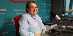 Na zdjęciu znajduje się lekko uśmiechnięta kobieta w okularach, która siedzi przed mikrofonem w studiu radiowym. Zdjęcie robiona z ukosa.