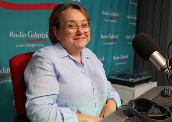 Na zdjęciu znajduje się lekko uśmiechnięta kobieta w okularach, która siedzi przed mikrofonem w studiu radiowym. Zdjęcie robiona z ukosa.