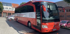 Czerwony autokar pełniący funkcję mobilnego punktu poboru krwi (krwiobus) Regionalnego Centrum Krwiodawstwa i Krwiolecznictwa w Gdańsku. Pojazd zaparkowany jest na wyłożonym kostką placu przed budynkami Komendy Miejskiej Policji w Sopocie. Na boku autokaru widnieje napis "Mobilny punkt poboru krwi" oraz logo Ministerstwa Zdrowia, a z przodu umieszczono symbol kropli krwi w kształcie serca. W tle widać fragmenty budynków oraz zaparkowane samochody osobowe