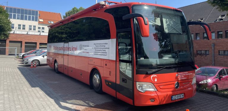Czerwony autokar pełniący funkcję mobilnego punktu poboru krwi (krwiobus) Regionalnego Centrum Krwiodawstwa i Krwiolecznictwa w Gdańsku. Pojazd zaparkowany jest na wyłożonym kostką placu przed budynkami Komendy Miejskiej Policji w Sopocie. Na boku autokaru widnieje napis "Mobilny punkt poboru krwi" oraz logo Ministerstwa Zdrowia, a z przodu umieszczono symbol kropli krwi w kształcie serca. W tle widać fragmenty budynków oraz zaparkowane samochody osobowe