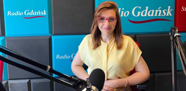 Monika Rapacewicz, rzeczniczka słupskiego magistratu, w okularach i żółtej bluzce siedząca przed mikrofonem w studiu Radia Gdańsk. Opowiada o wakacyjnych remontach szkół i przedszkoli. W tle widoczne są niebiesko-czarne panele akustyczne z logo stacji