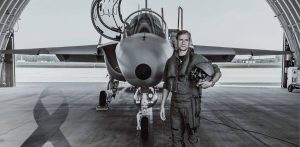 Zdjęcie czarno-białe. Major pilot Robert Jeł stoi przed samolotem szkolno-treningowym M-346 Bielik w hangarze, trzymając w ręku kask lotniczy. W dolnym lewym rogu widoczna jest czarna żałobna wstęga. Żołnierz zginął 12 lipca 2024 roku w katastrofie samolotu M-346 Bielik na terenie 43 Bazy Lotnictwa. Wydział ds. Wojskowych Prokuratury Okręgowej w Gdańsku poinformował o umorzeniu śledztwa w tej sprawie
