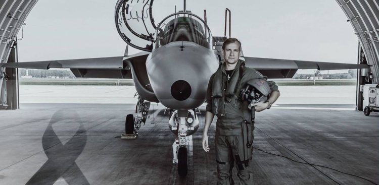 Zdjęcie czarno-białe. Major pilot Robert Jeł stoi przed samolotem szkolno-treningowym M-346 Bielik w hangarze, trzymając w ręku kask lotniczy. W dolnym lewym rogu widoczna jest czarna żałobna wstęga. Żołnierz zginął 12 lipca 2024 roku w katastrofie samolotu M-346 Bielik na terenie 43 Bazy Lotnictwa. Wydział ds. Wojskowych Prokuratury Okręgowej w Gdańsku poinformował o umorzeniu śledztwa w tej sprawie