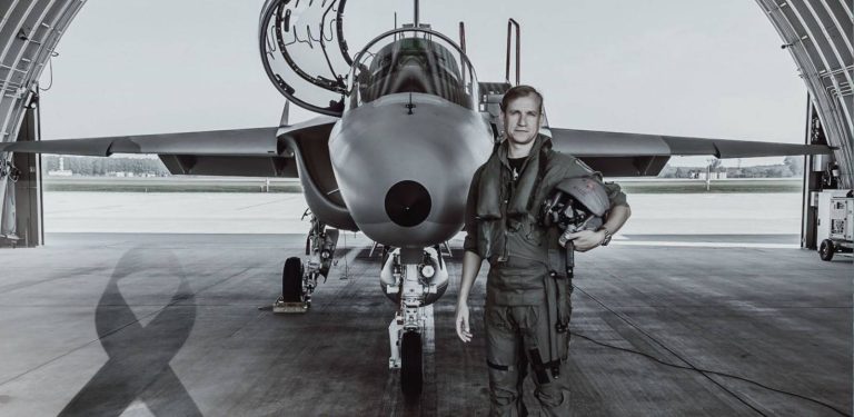 Zdjęcie czarno-białe. Major pilot Robert Jeł stoi przed samolotem szkolno-treningowym M-346 Bielik w hangarze, trzymając w ręku kask lotniczy. W dolnym lewym rogu widoczna jest czarna żałobna wstęga. Żołnierz zginął 12 lipca 2024 roku w katastrofie samolotu M-346 Bielik na terenie 43 Bazy Lotnictwa. Wydział ds. Wojskowych Prokuratury Okręgowej w Gdańsku poinformował o umorzeniu śledztwa w tej sprawie