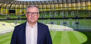 Jacek Jakubowski, nowy dyrektor generalny Lechii Gdańsk, w okularach, białej koszuli i granatowej marynarce, pozuje na tle pustych, zielono-żółtych trybun stadionu w Gdańsku