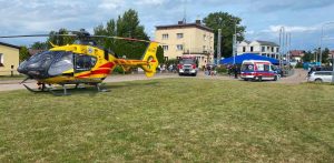 Akcja ratunkowa po wybuchu na kutrze w Jastarni. Żółto-czerwony śmigłowiec Lotniczego Pogotowia Ratunkowego (LPR) stojący na trawniku w terenie miejskim. W tle widoczna jest karetka pogotowia, wóz strażacki oraz budynki