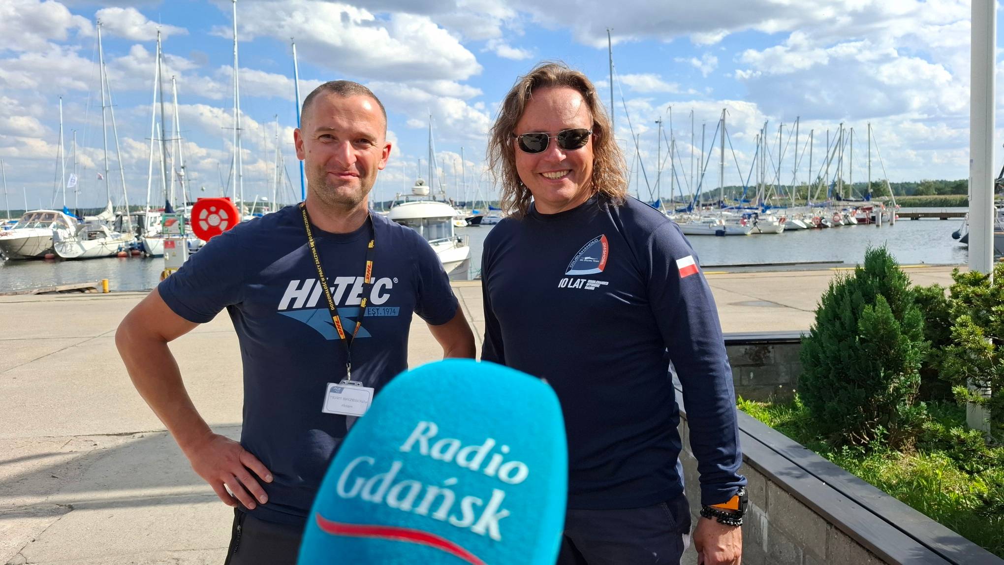 Dwaj mężczyźni w sportowych koszulkach stoją na nabrzeżu w marinie, na pierwszym planie mikrofon Radia Gdańsk