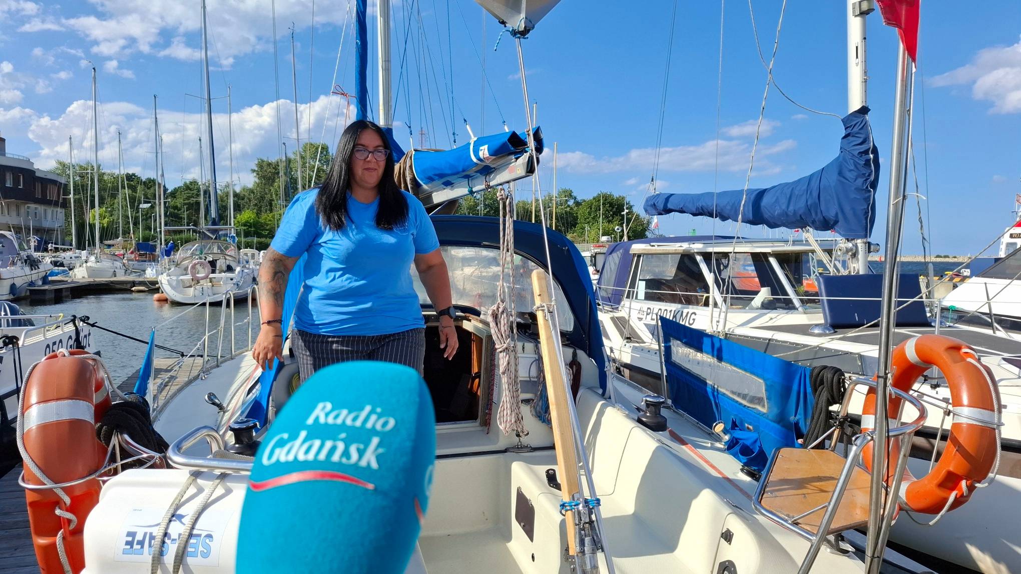 Kobieta w niebieskiej koszulce stojąca na pokładzie jachtu w marinie, na pierwszym planie mikrofon Radia Gdańsk