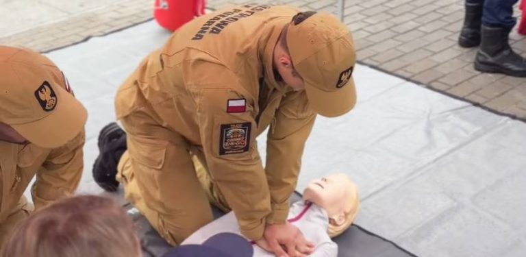 Dwóch strażaków w beżowych mundurach i czapeczkach z polską flagą na ramieniu prowadzi pokaz pierwszej pomocy na manekinie treningowym na świeżym powietrzu. Zajęcia w ramach Krajowego Treningu Reagowania Akademii Ochrony Ludności i Obrony Cywilnej przy Plaży Śródmieście w Gdyni