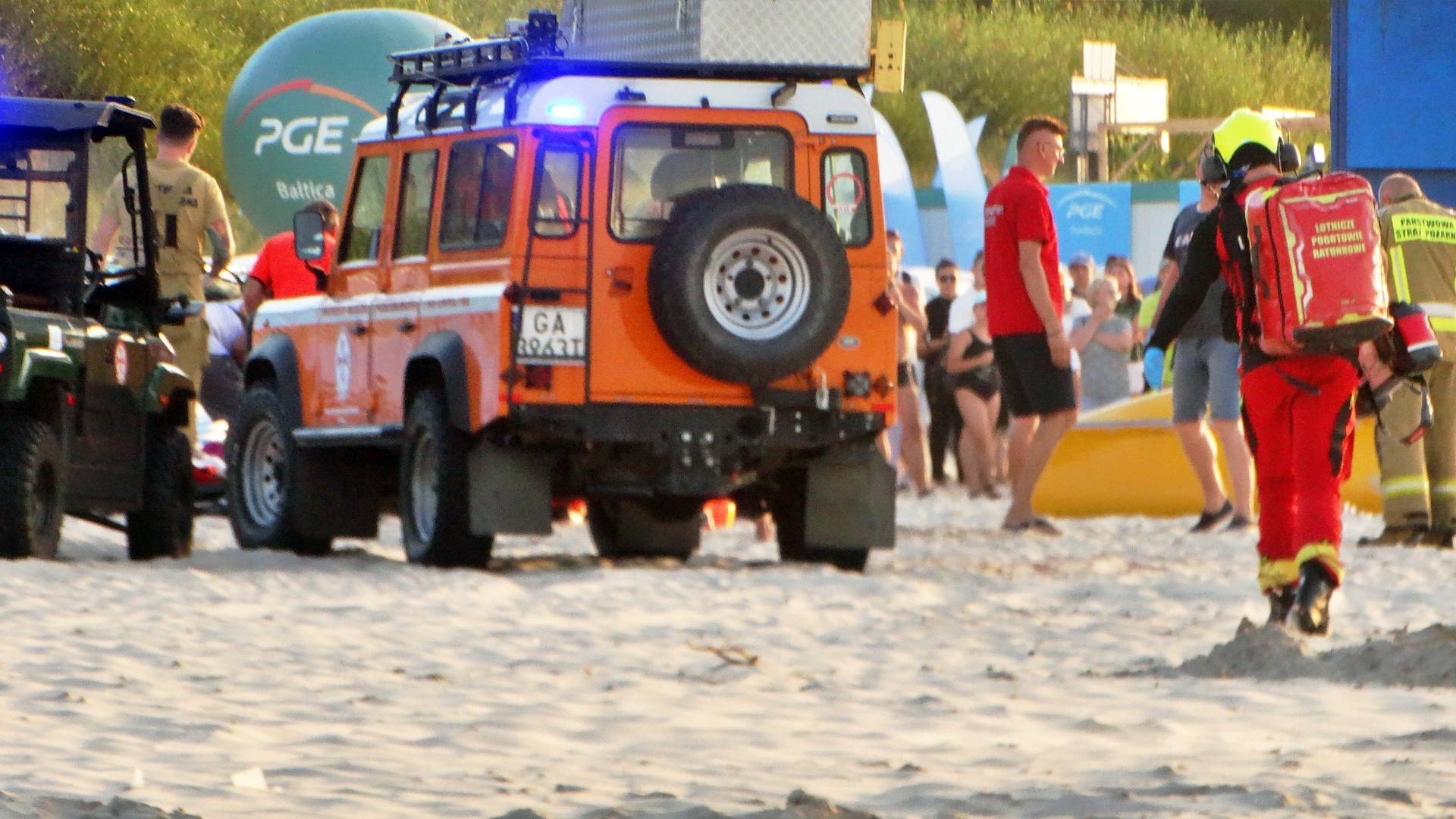 Służby ratunkowe i tłum na plaży w Ustce podczas wieczornej interwencji po wyciągnięciu z morza nietrzeźwej kobiety. Widoczny ratownik medyczny, strażak i pojazd terenowy