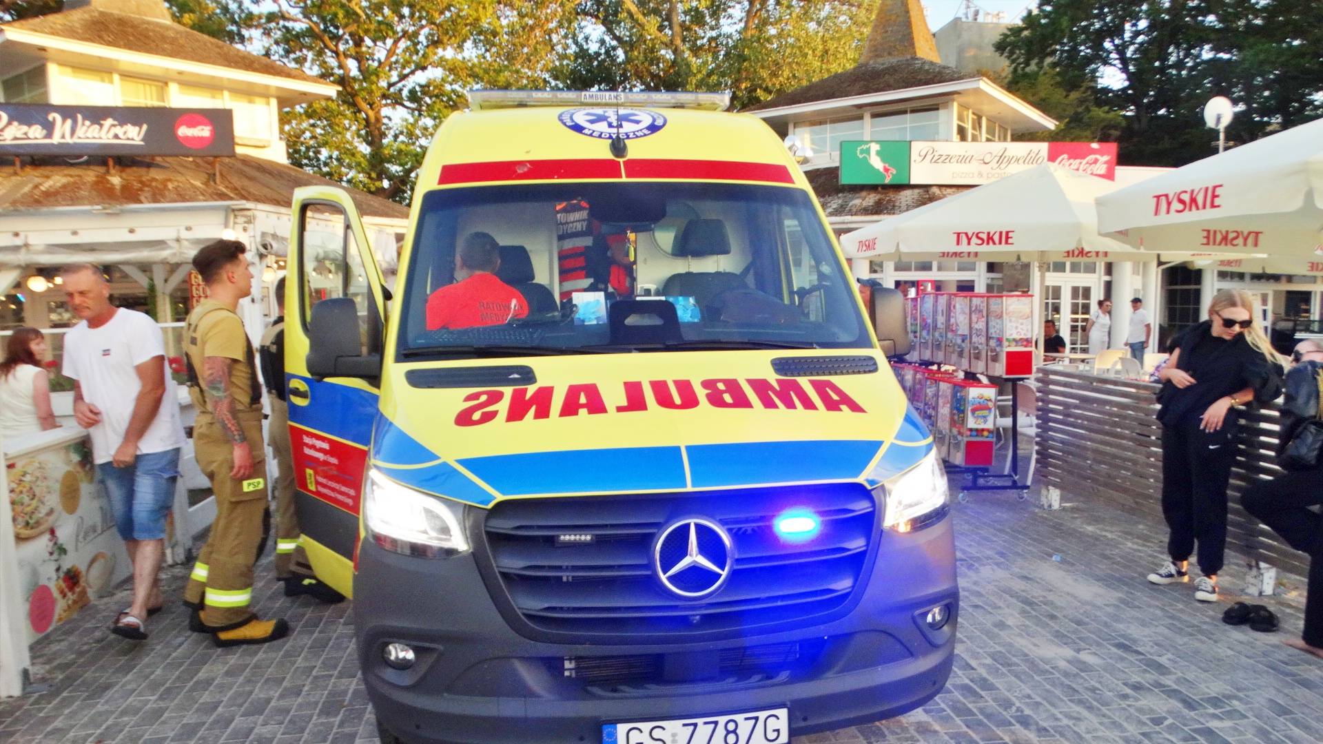 Żółty ambulans z włączonym sygnałem świetlnym zaparkowany na promenadzie w Ustce podczas akcji ratunkowej, obok strażaka i przechodniów