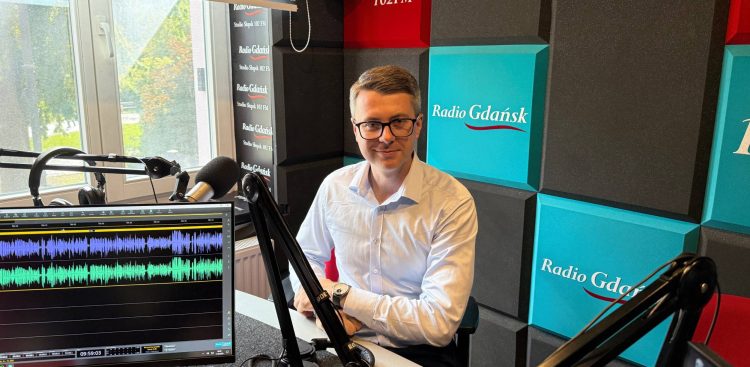 Mężczyzna w błękitnej koszuli i okularach, europoseł Piotr Müller, siedzi przy mikrofonie w studiu nagraniowym Radia Gdańsk w Słupsku. Na pierwszym planie widać monitor z otwartym programem do edycji dźwięku pokazującym fale dźwiękowe, a w tle ścianę z logo Radia Gdańsk