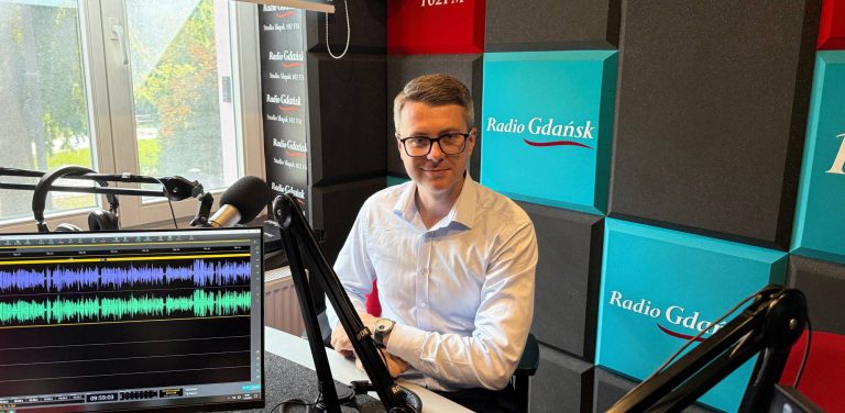 Mężczyzna w błękitnej koszuli i okularach, europoseł Piotr Müller, siedzi przy mikrofonie w studiu nagraniowym Radia Gdańsk w Słupsku. Na pierwszym planie widać monitor z otwartym programem do edycji dźwięku pokazującym fale dźwiękowe, a w tle ścianę z logo Radia Gdańsk