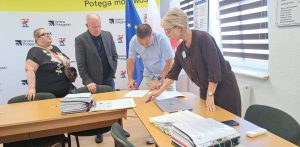 Cztery osoby stoją przy stole w biurze i pochylają się nad dokumentami. Kobieta i mężczyzna w centrum podpisują oraz sprawdzają papiery, obok leżą grube teczki z dokumentacją. W tle widoczne są flagi Polski i Unii Europejskiej oraz ścianka z logo Gminy Potęgowo, w której powstaną nowe mieszkania socjalne