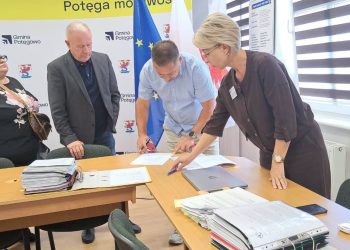 Cztery osoby stoją przy stole w biurze i pochylają się nad dokumentami. Kobieta i mężczyzna w centrum podpisują oraz sprawdzają papiery, obok leżą grube teczki z dokumentacją. W tle widoczne są flagi Polski i Unii Europejskiej oraz ścianka z logo Gminy Potęgowo, w której powstaną nowe mieszkania socjalne
