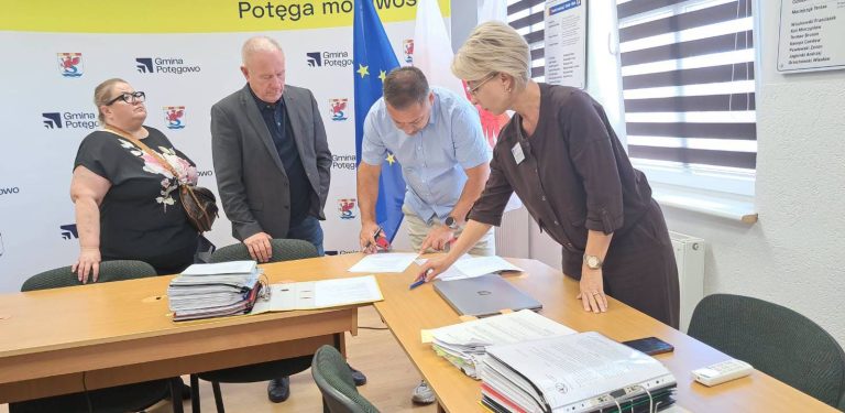 Cztery osoby stoją przy stole w biurze i pochylają się nad dokumentami. Kobieta i mężczyzna w centrum podpisują oraz sprawdzają papiery, obok leżą grube teczki z dokumentacją. W tle widoczne są flagi Polski i Unii Europejskiej oraz ścianka z logo Gminy Potęgowo, w której powstaną nowe mieszkania socjalne