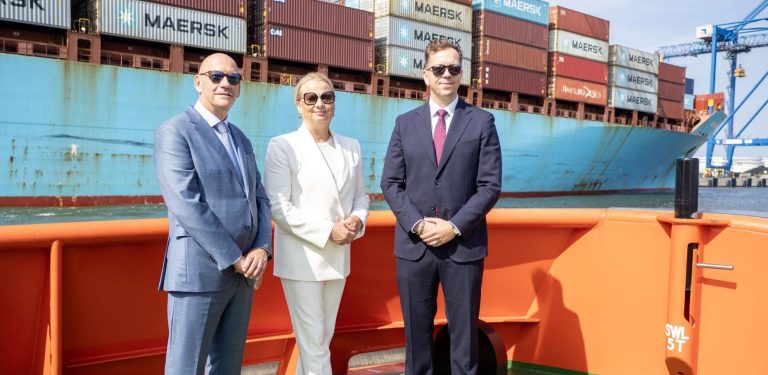 Trzy osoby w garniturach i okularach przeciwsłonecznych stoją na tle dużego kontenerowca Maersk oraz suwnic w porcie w Gdańsku