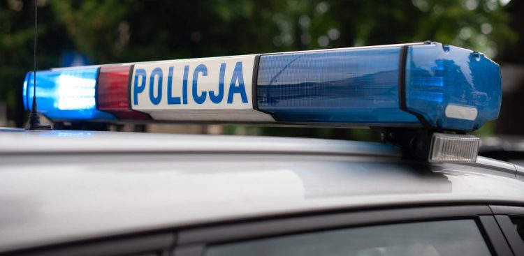 Zbliżenie na belkę sygnalizacyjną na dachu radiowozu policyjnego z widocznym napisem "policja" oraz włączonymi niebieskimi i czerwonymi światłami. Policja apeluje o trzeźwość do kierowców na drodze wojewódzkiej 502 na Mierzeję Wiślaną