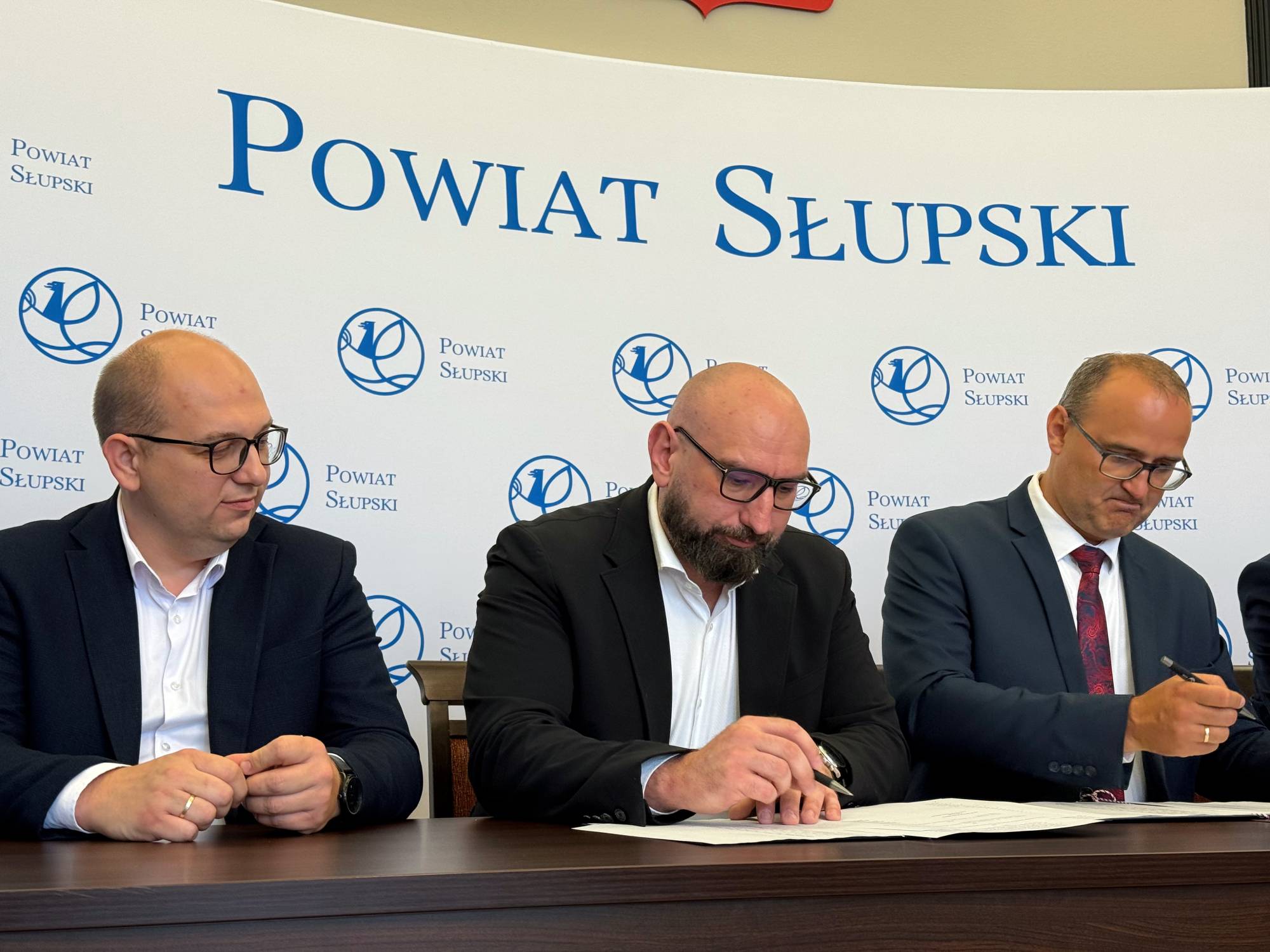 Wicewojewoda pomorski Emil Rojek, wicestarosta Marcin Kowalczyk oraz starosta słupski Paweł Lisowski podczas podpisywania umowy na tle ścianki z napisem Powiat Słupski