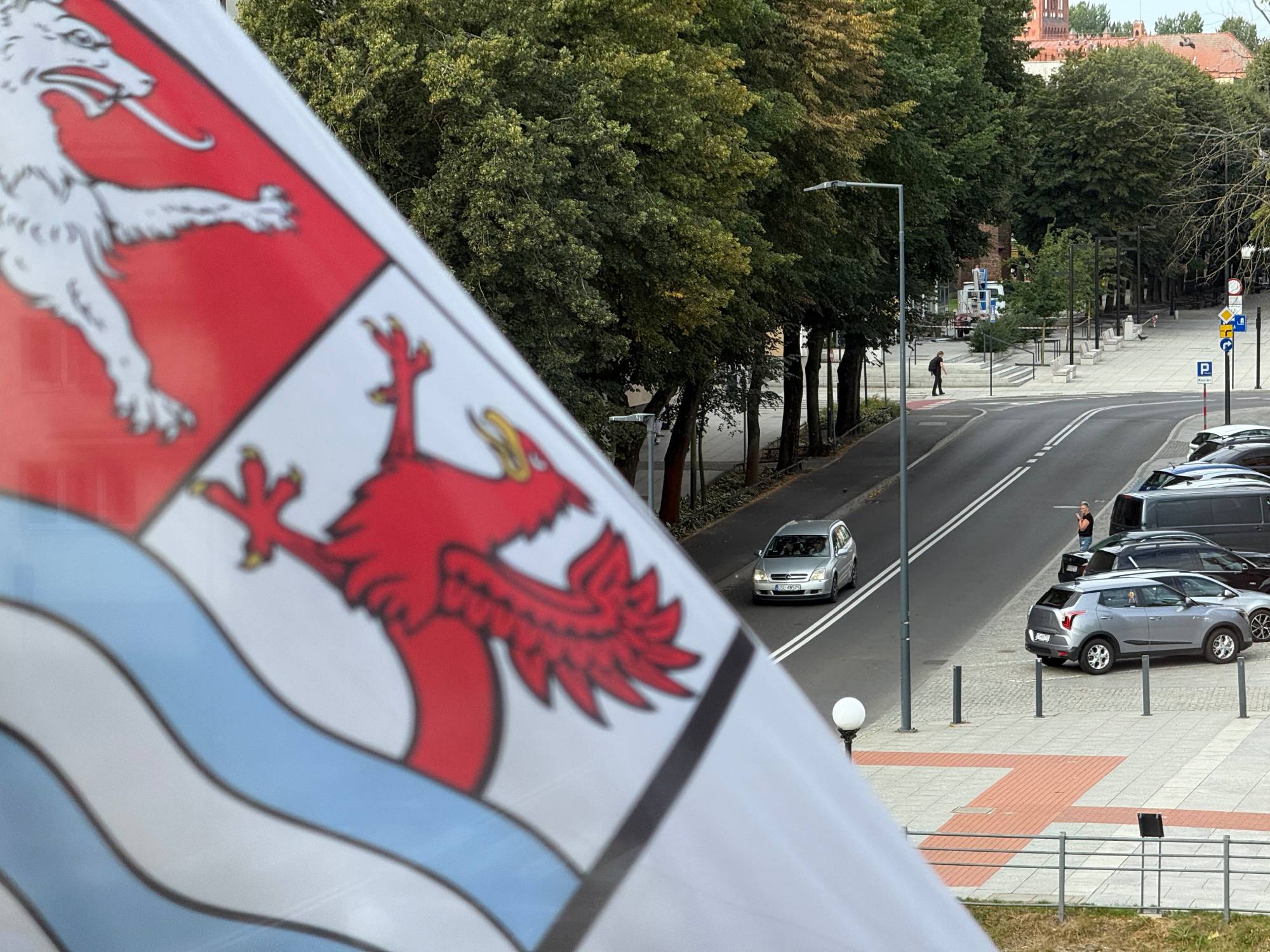 Widok z góry na miejską ulicę z jadącym samochodem, z powiewającą na pierwszym planie flagą z herbem powiatu słupskiego