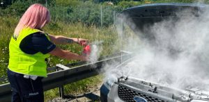 Pożar samochodu na trasie S6. Policjantka z różowymi włosami w odblaskowej kamizelce gasi gaśnicą samochodową dymiący silnik pod otwartą maską srebrnego forda stojącego przy barierze energochłonnej.