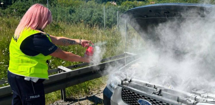 Pożar samochodu na trasie S6. Policjantka z różowymi włosami w odblaskowej kamizelce gasi gaśnicą samochodową dymiący silnik pod otwartą maską srebrnego forda stojącego przy barierze energochłonnej.