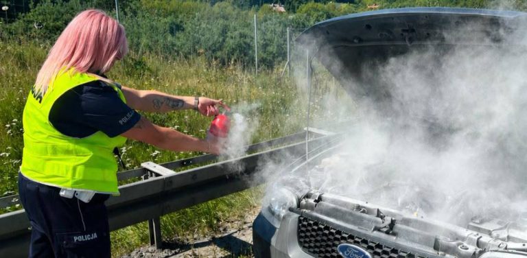 Pożar samochodu na trasie S6. Policjantka z różowymi włosami w odblaskowej kamizelce gasi gaśnicą samochodową dymiący silnik pod otwartą maską srebrnego forda stojącego przy barierze energochłonnej.