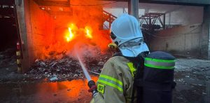 Pożar w Chlewnicy. Strażak z Ochotniczej Straży Pożarnej w hełmie i z aparatem oddechowym na plecach, widziany od tyłu, kieruje strumień wody z węża na płonącą stertę odpadów w hali