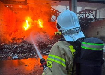 Pożar w Chlewnicy. Strażak z Ochotniczej Straży Pożarnej w hełmie i z aparatem oddechowym na plecach, widziany od tyłu, kieruje strumień wody z węża na płonącą stertę odpadów w hali